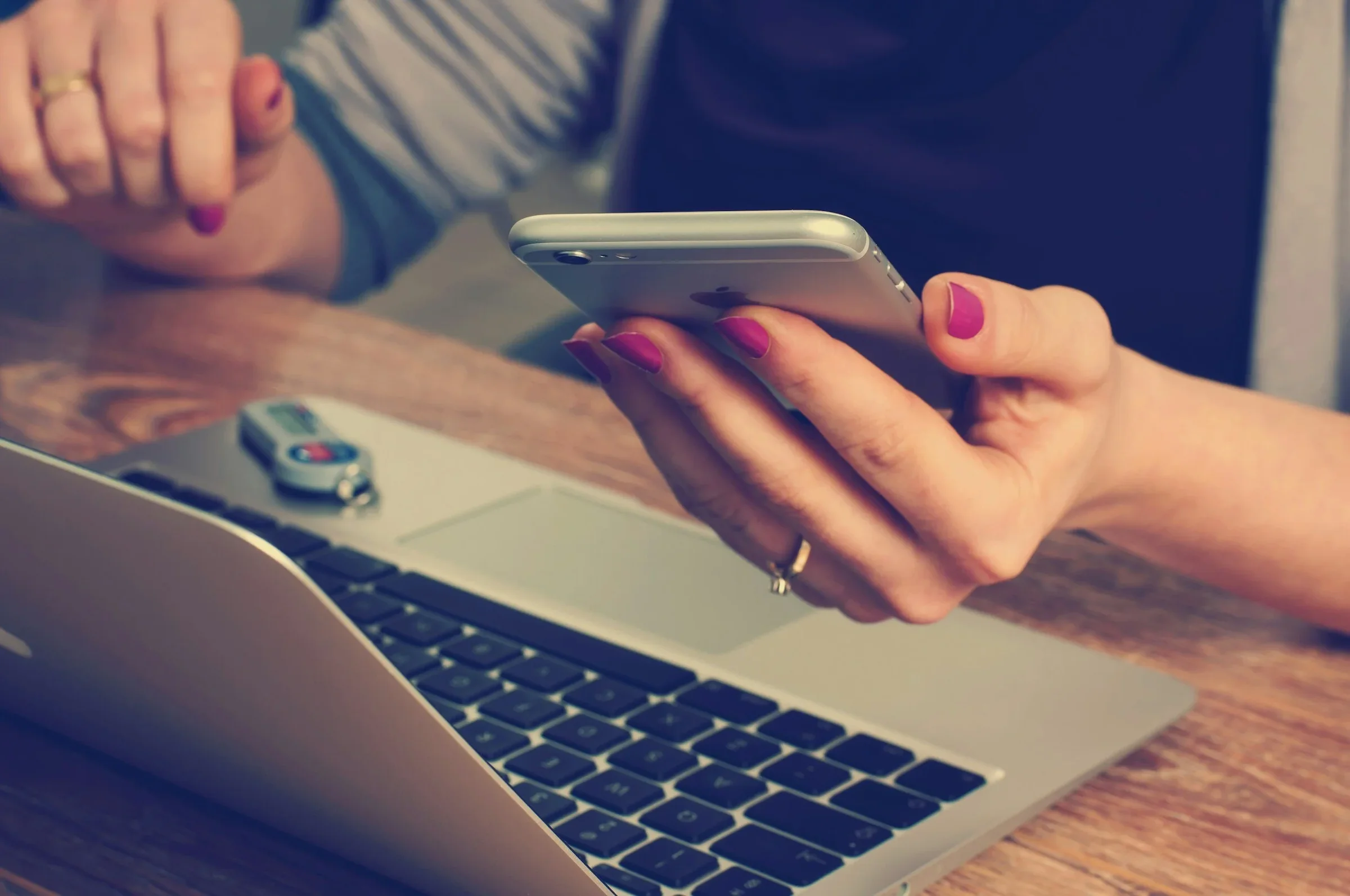 A person with pink painted nails holding a smartphone above a laptop on a wooden desk