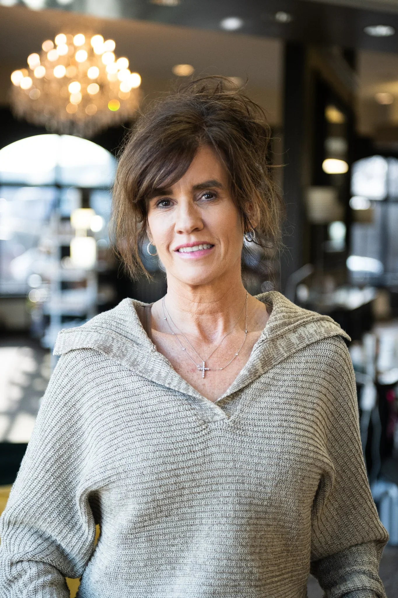 A woman with short brown hair and earrings smiling, wearing a beige knit sweater