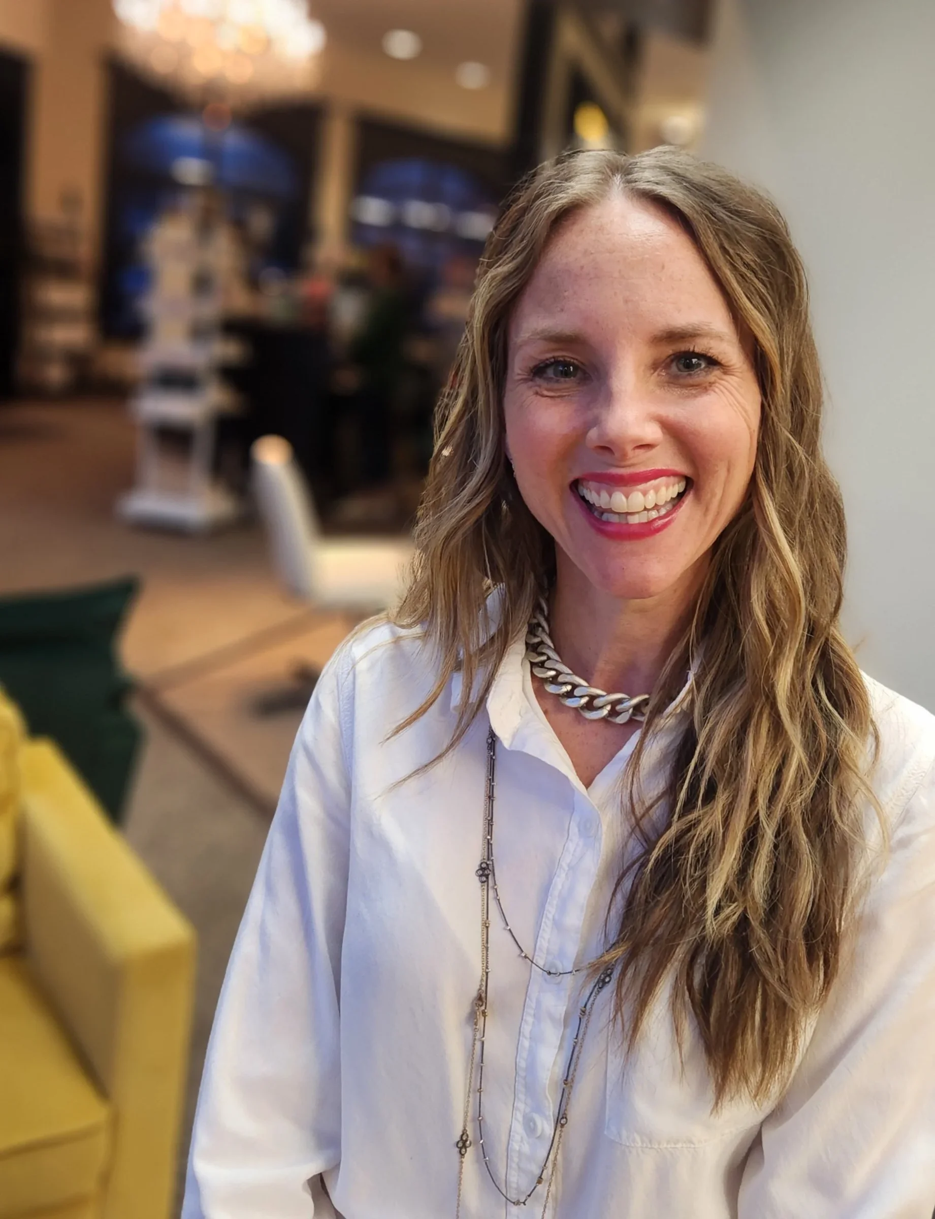 A woman with long wavy hair, wearing a white shirt, and layered necklaces, smiling in a warmly lit indoor setting.