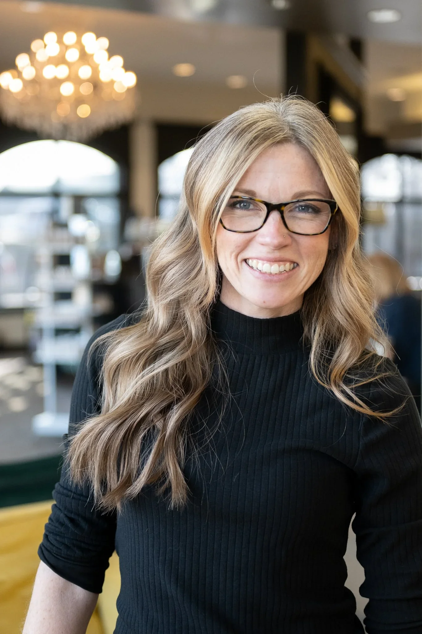 A woman with long wavy blonde hair and glasses smiling, wearing a black ribbed turtleneck