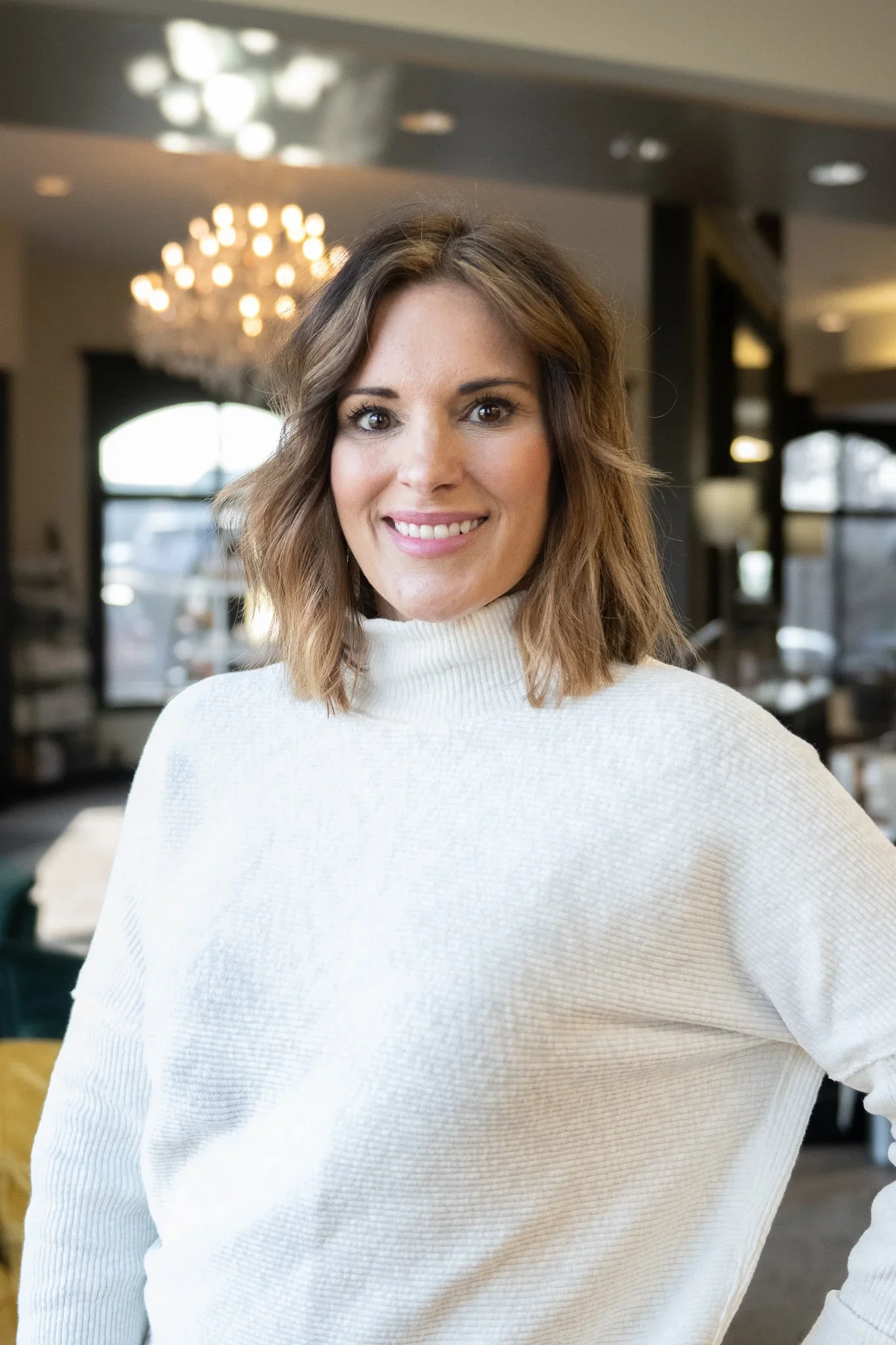 A woman with shoulder-length wavy brown hair, wearing a white turtleneck sweater