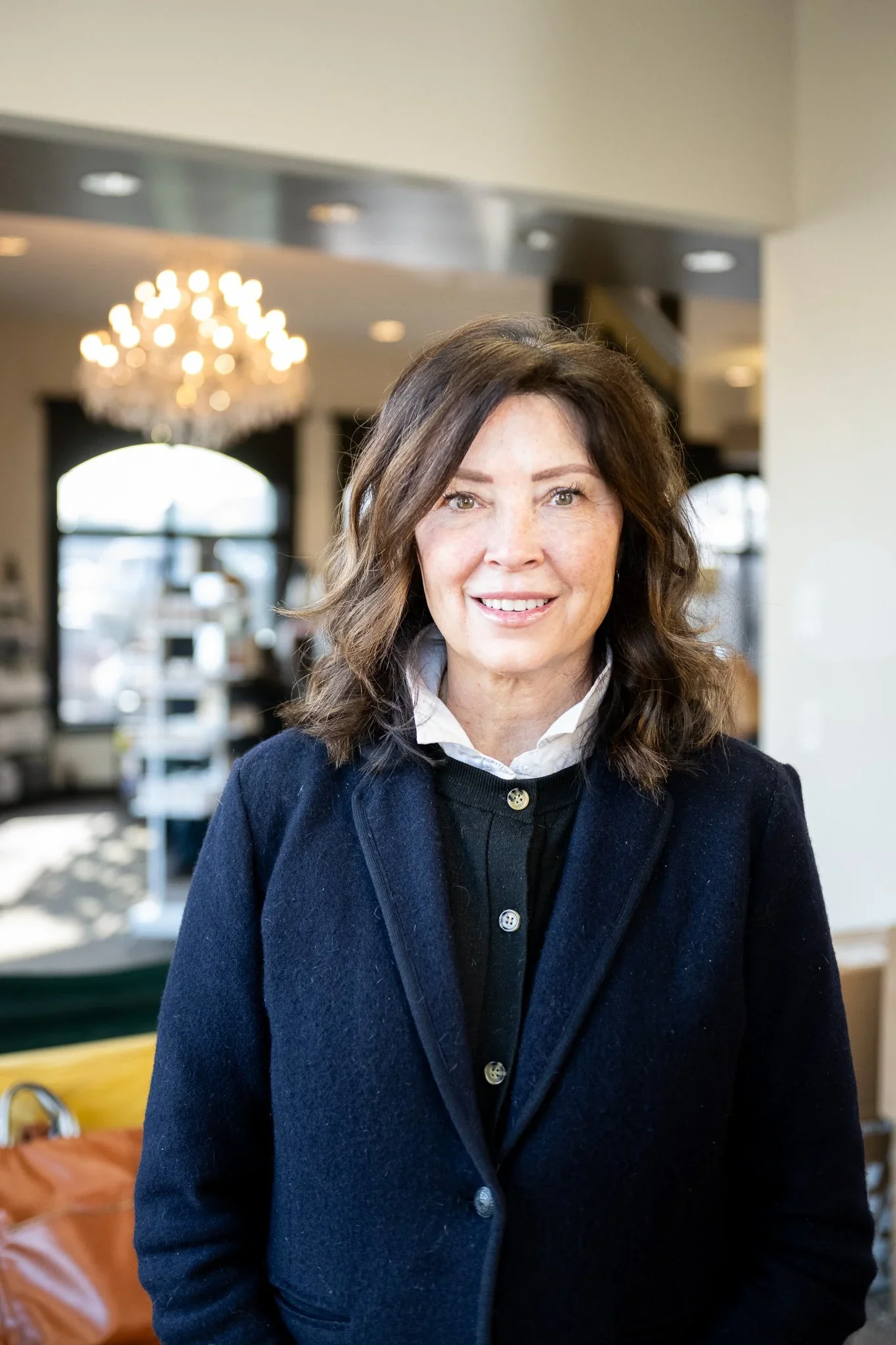 A woman with shoulder-length brown hair, wearing a dark blue coat over a white collared shirt