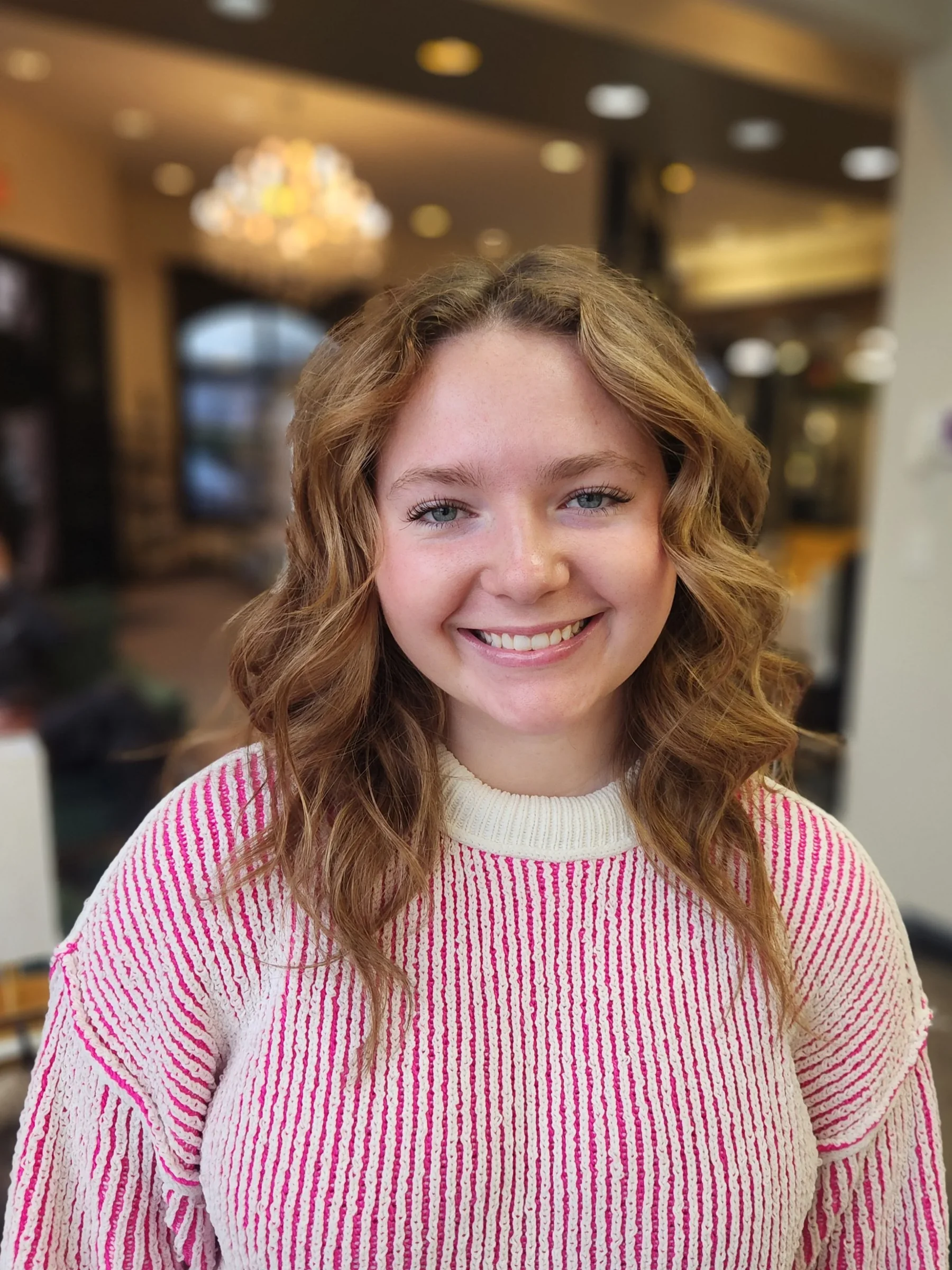 A smiling young woman with wavy, shoulder-length hair wearing a pink and white striped sweater