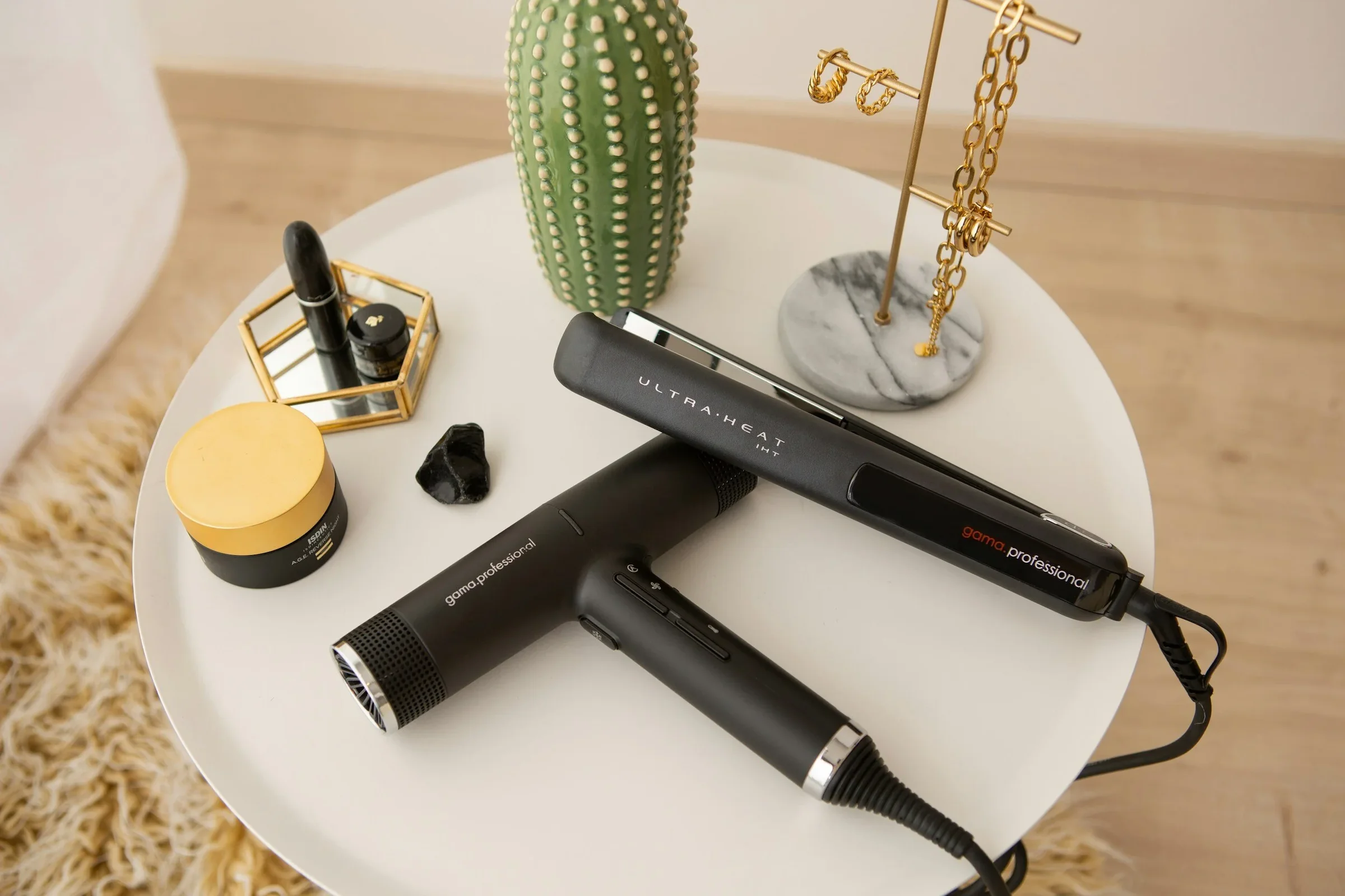A white round table holding a black professional hair dryer, a black and silver hair straightener, and a gold jewelry stand with chains, earrings, and a ring