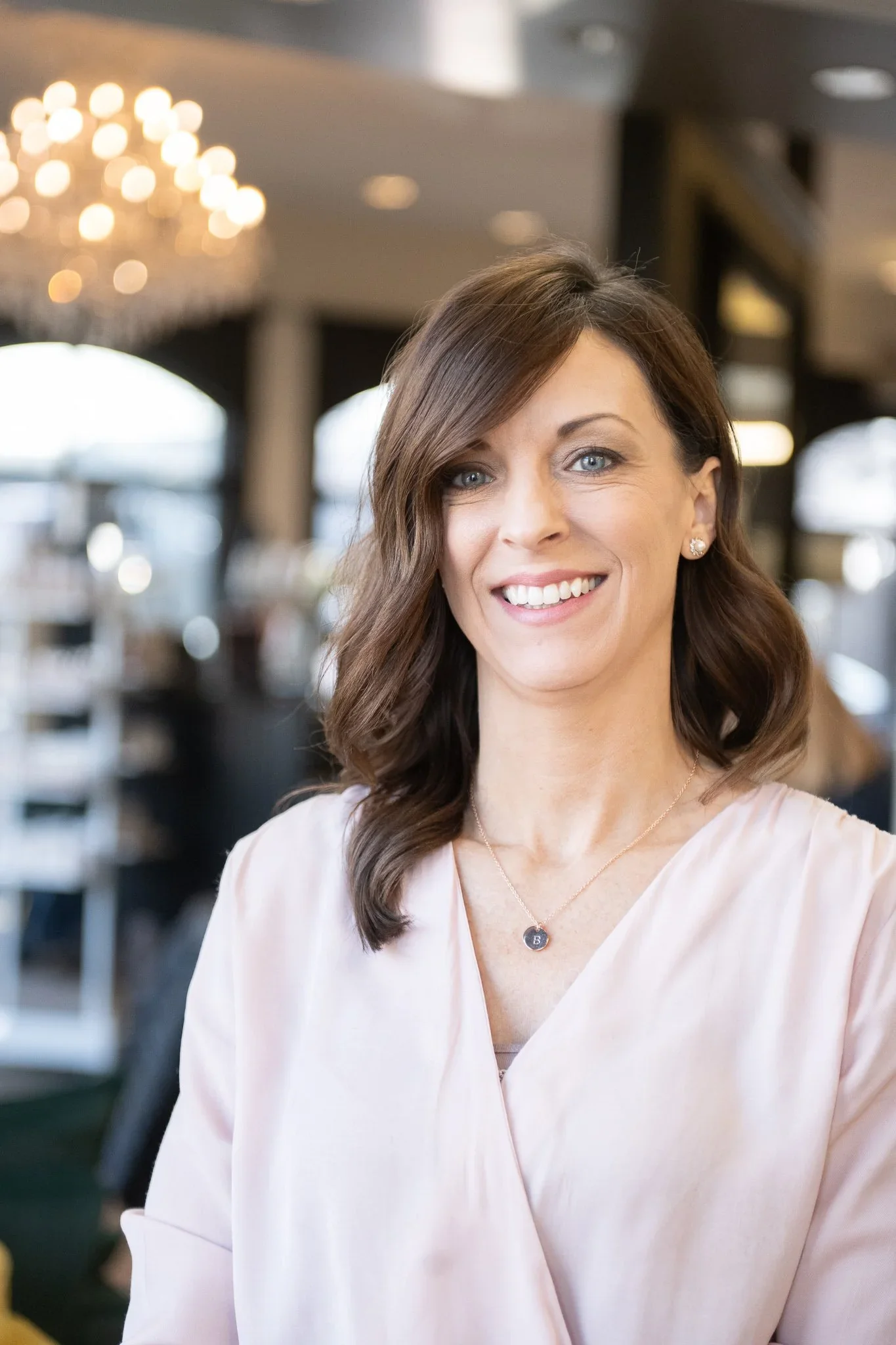 A woman with shoulder-length brown hair and blue eyes, smiling, wearing a pale pink blouse, pearl earrings, and a necklace with a pendant