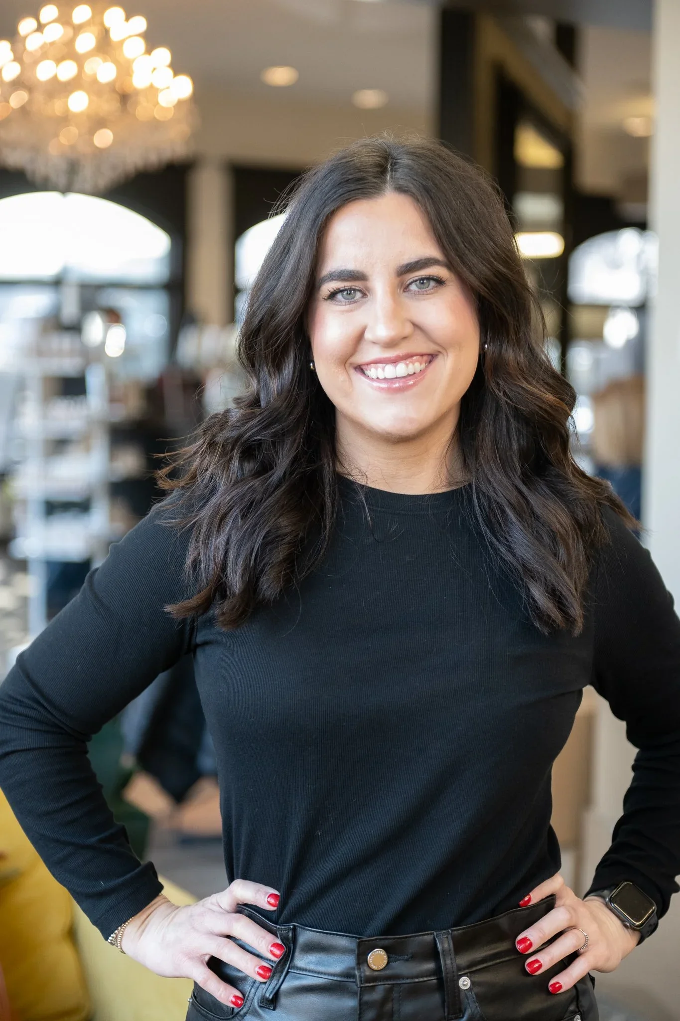 A woman with dark wavy hair smiling, wearing a black long-sleeve top and black leather pants, standing indoors