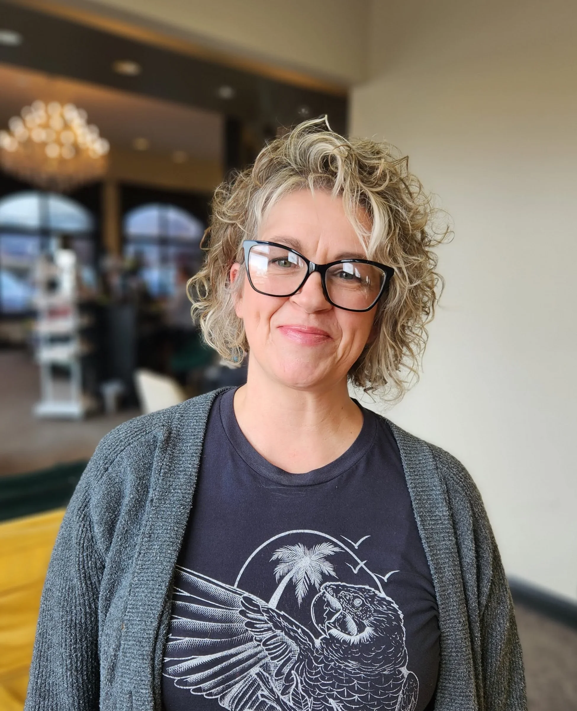 A woman with curly blonde hair and glasses smiling in an indoor setting