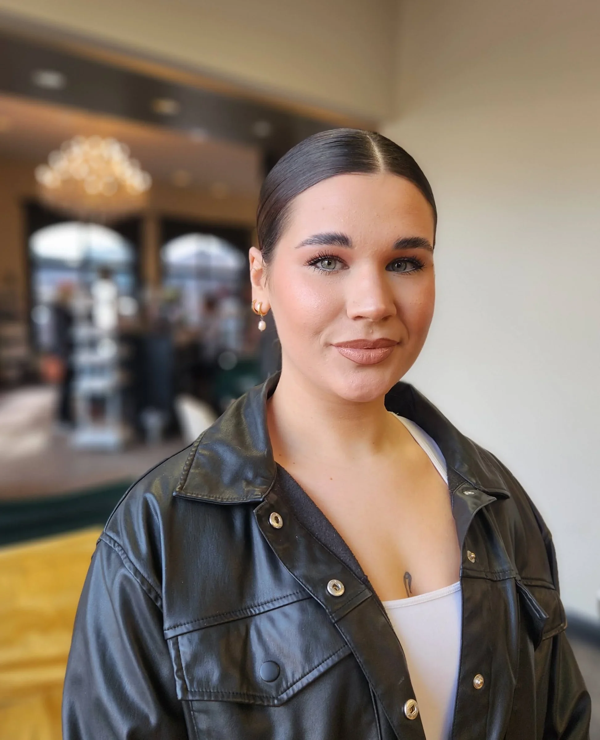A young woman with dark hair, wearing a black leather jacket, standing indoors