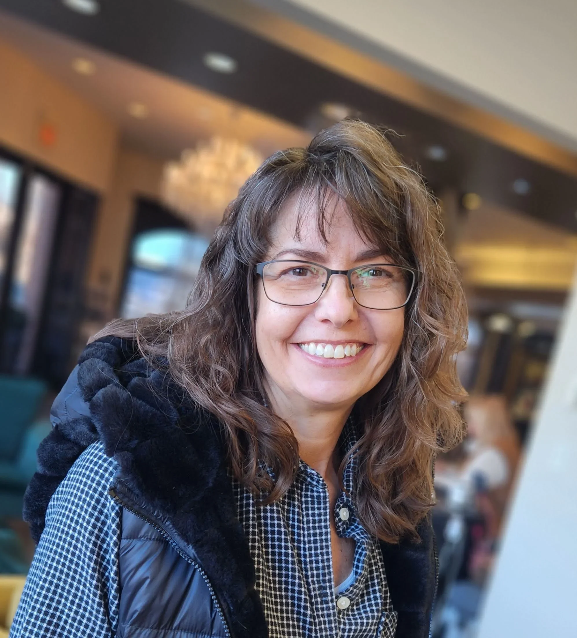 A woman with brown, curly hair and glasses smiles at the camera inside a well-lit, elegant indoor space.