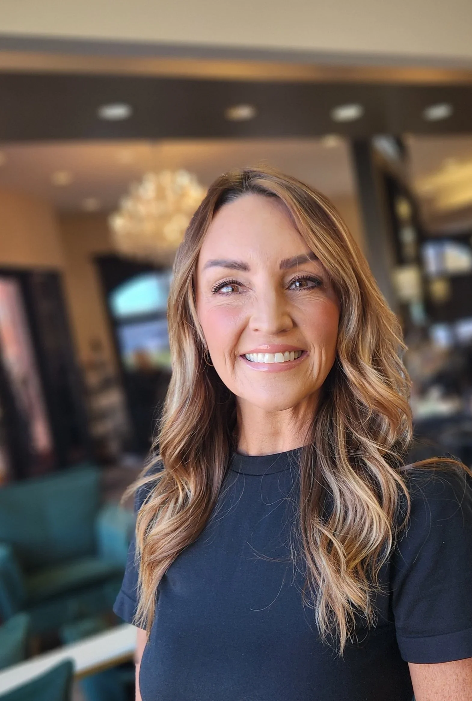 A woman with long wavy brown hair and a bright smile in a cheerful indoor setting